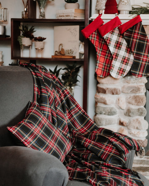 A red, black, and green plaid throw blanket and matching pillow are draped over a gray armchair in a cozy living room setting, with matching plaid Christmas stockings hanging on a stone fireplace mantel in the background.
