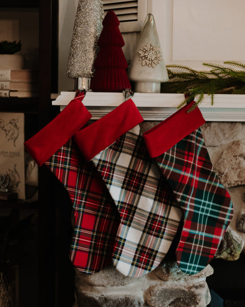Three Christmas stockings with red cuffs hang on a stone fireplace mantel. The stockings feature red, green, and white plaid patterns, with decorative holiday trees and greenery on the mantel above.