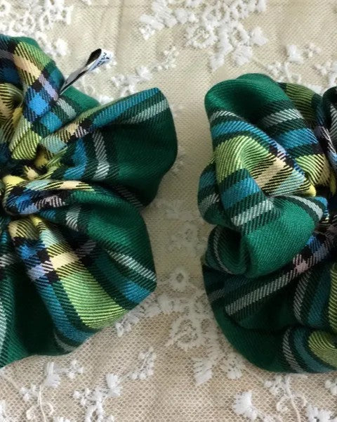 Two green blue yellow and white plaid hair scrunchies on a textured beige surface.