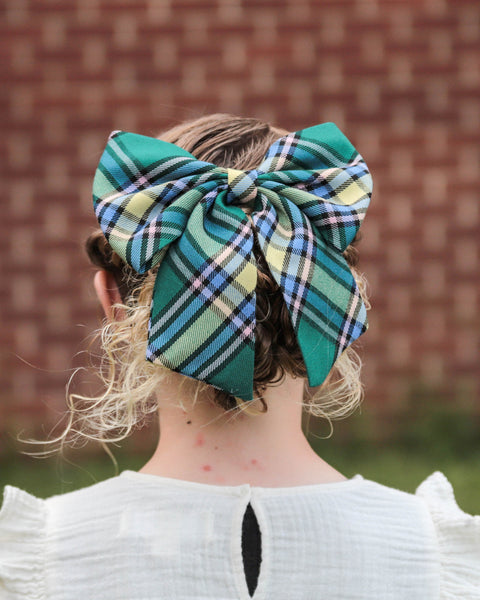 Girl wearing a large green and yellow plaid bow in their hair with a brick wall background