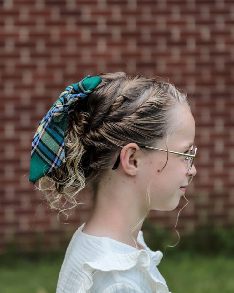 Girl with braided hair and plaid hair bow looking away from the camera in front of a brick wall