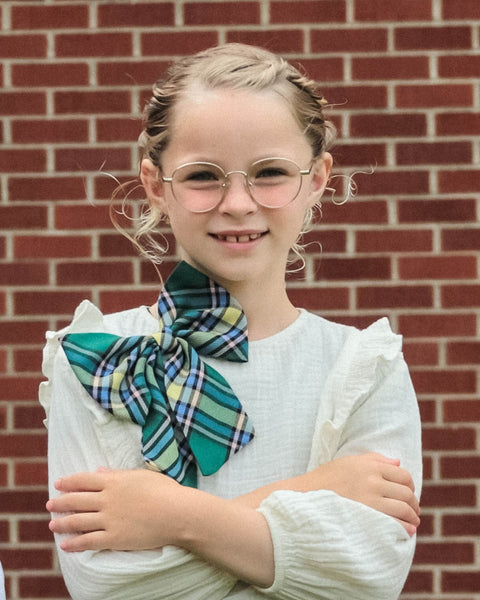 Young girl wearing a plaid neck bow and pink glasses in front of a brick wall