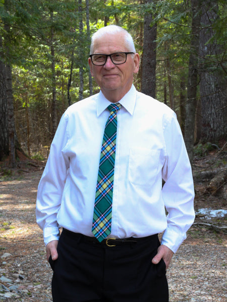 Senior man wearing a white shirt, black pants, and a green plaid tie standing in a forest.