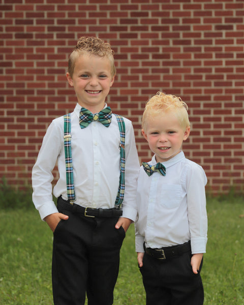 Two young boys wearing white shirts, black pants, and Alberta tartan plaid bow ties standing in front of a brick wall.