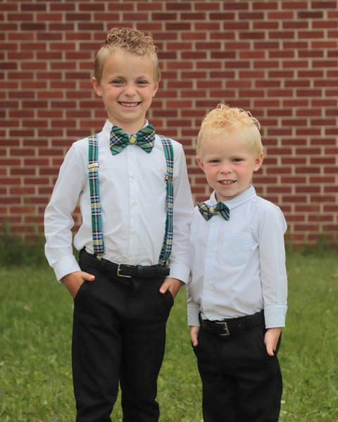 Two young boys wiht curly hair in formal attire with green plaid suspenders and bow ties standing in front of a brick wall.