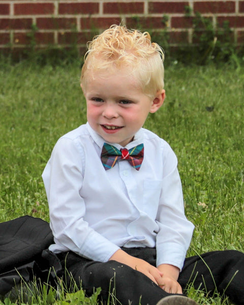 Child wearing a white shirt and British Columbia plaid bow tie sitting on grass with a brick wall in the background