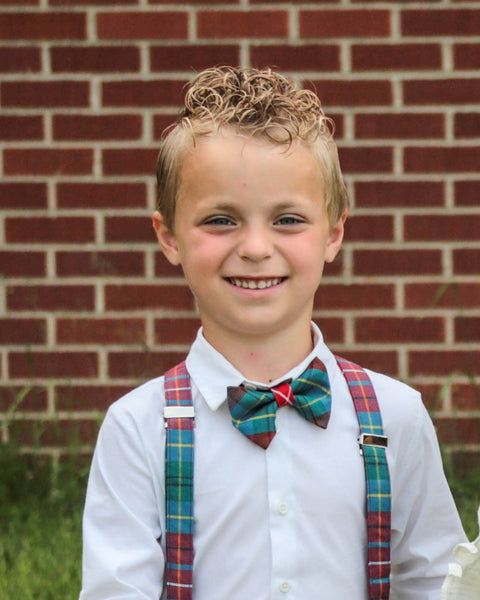 Young boy wearing a British Columbia tartan plaid bow tie and suspenders in front of a brick wall.
