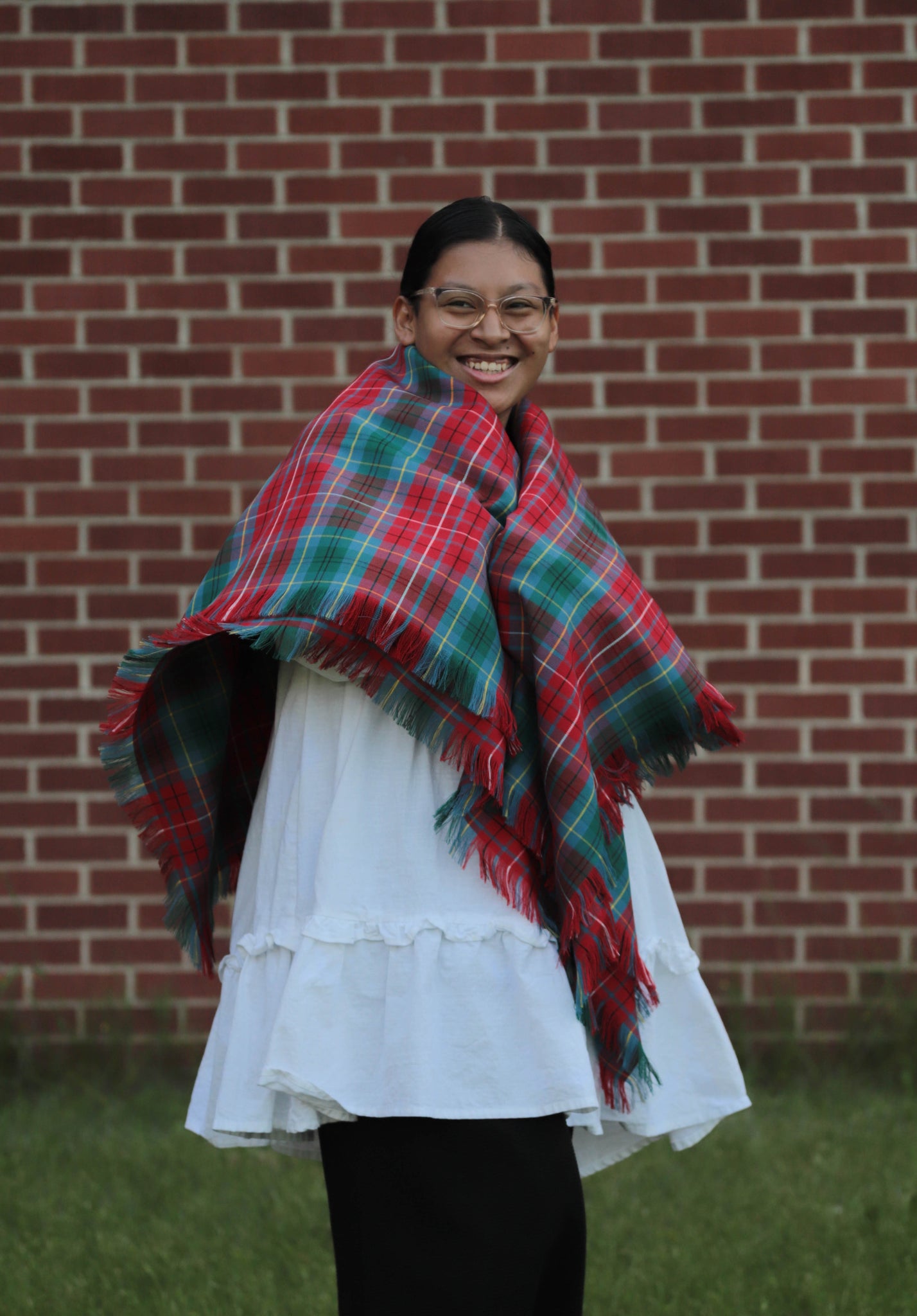Person wearing a British Columbia tartan red and green plaid blanket scarf shawl in front of a brick wall.