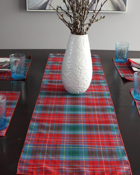 Dark dining table set with a red and blue plaid runner, white vase with pussy willows, and blue glasses.