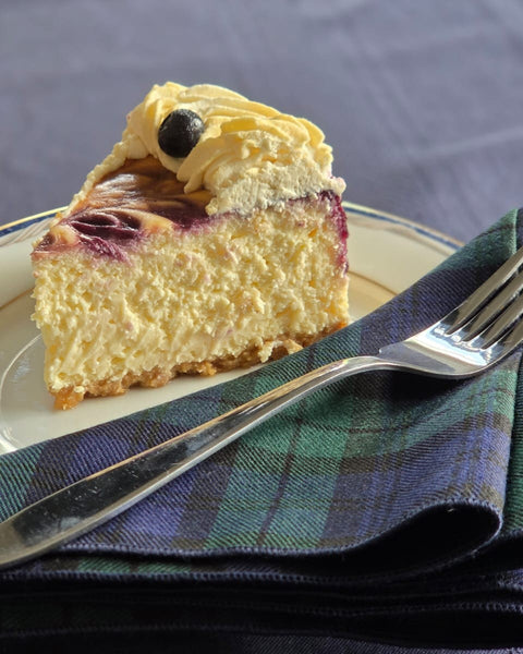 Slice of cheesecake with blueberries on a plate with a fork, on a navy and black plaid napkin.