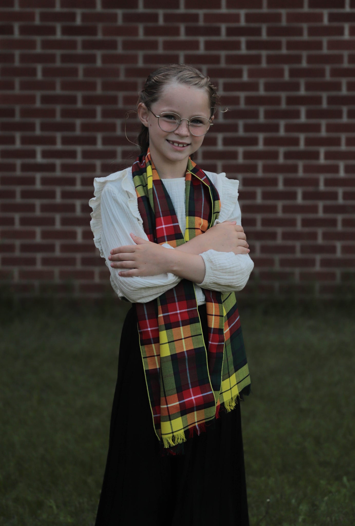 Young girl wearing a Buchanan tartan yellow, red and white plaid scarf over a white blouse, standing in front of a brick wall with her arms crossed over the scarf.