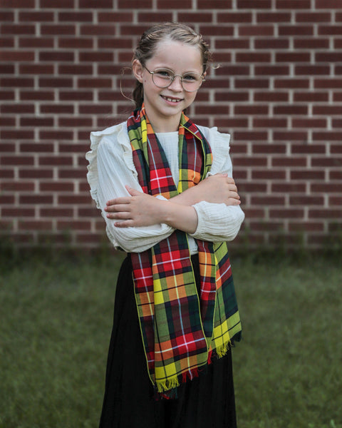 Young girl wearing abright yellow, red, green plaid scarf in front of a brick wall