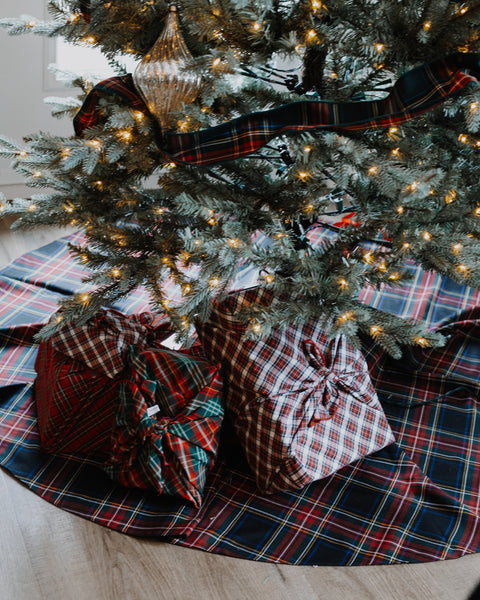 A festive Christmas tree skirt in a red, green, and navy tartan plaid pattern, styled under a decorated Christmas tree with warm lights and wrapped presents on a light wood floor.