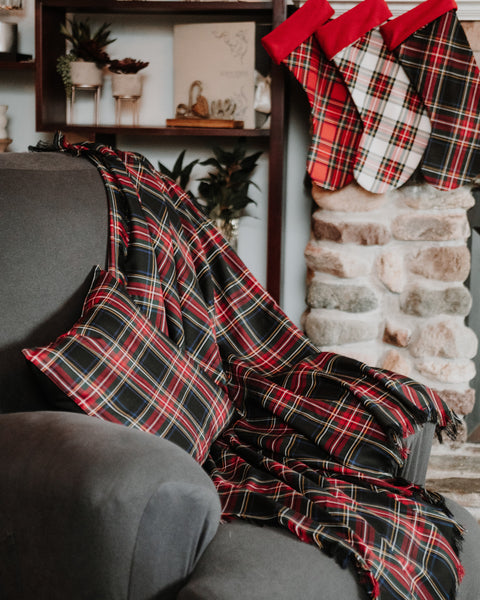 A red, black, and white plaid Black Stewart tartan throw blanket is draped over a gray armchair in a cozy living room setting, with matching plaid stockings hanging on a stone fireplace.