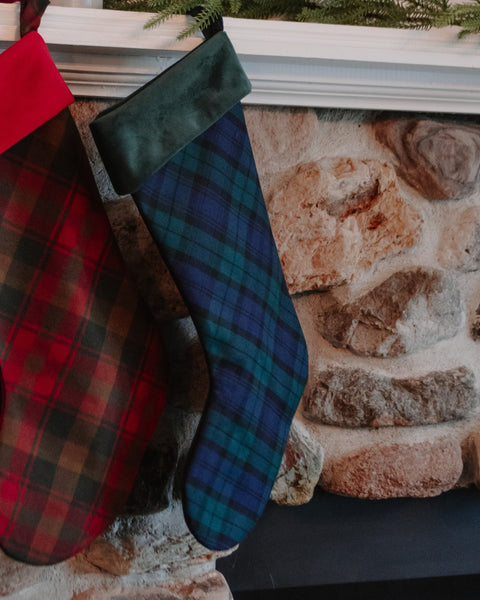 Two Christmas stockings hanging on a stone fireplace mantel. One stocking is red with a black and green plaid pattern, and the other is dark green velvet with a blue and green tartan plaid body.