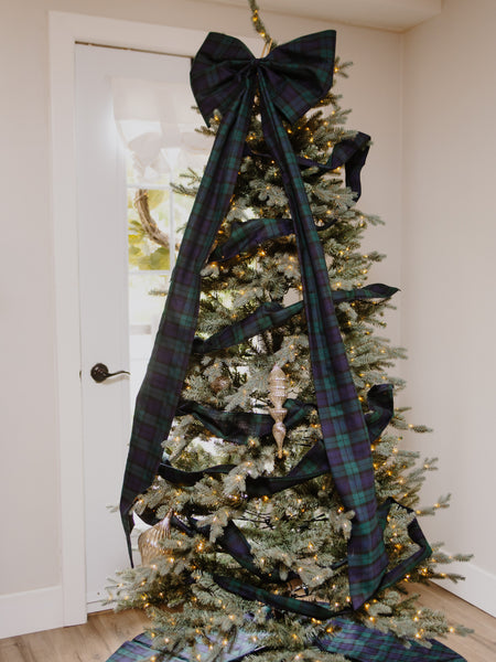 A decorated Christmas tree in a home interior, featuring a large dark green and black plaid Black Watch tartan ribbon bow at the top, with matching plaid ribbon draped down the tree and wrapped around it. The tree is lit with warm white string lights and has small ornaments, including a striped pinecone ornament.