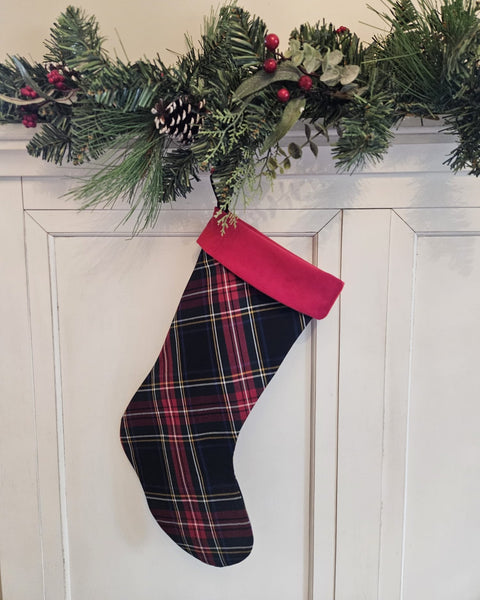 A Christmas stocking with a red and black plaid body and a bright red cuff, hanging from a green pine garland with red berries on a white paneled door.