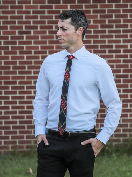 Man wearing a white dress shirt, black pants, and a black and red plaid tie standing against a brick wall.