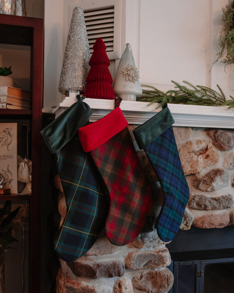 Three Christmas stockings with tartan plaid patterns hang on a stone fireplace mantel next to a dark wood shelf. The stockings feature green, red, and blue plaid designs with solid red and dark green cuffs. Black Watch tartan, Maple Leaf tartan, and Hunting Stewart tartan christmas stockings