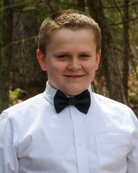 Young boy wearing a white shirt and Black Watch tartan bow tie outdoors with trees in the background