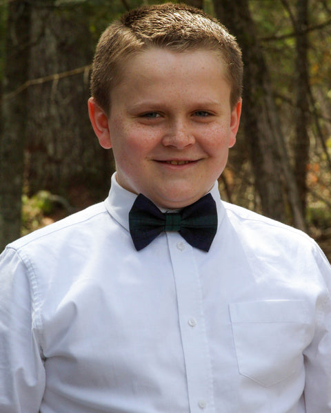 Young boy wearing a white shirt and black watch tartan bow tie outdoors with trees in the background