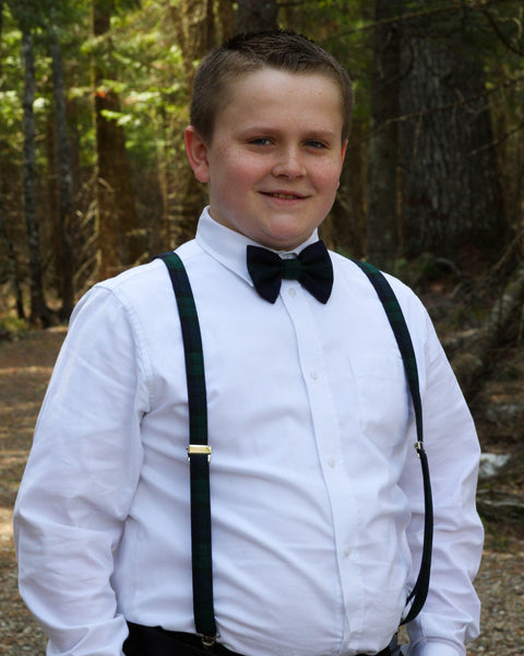 Young boy in formal attire with suspenders and bow tie standing in a forest.