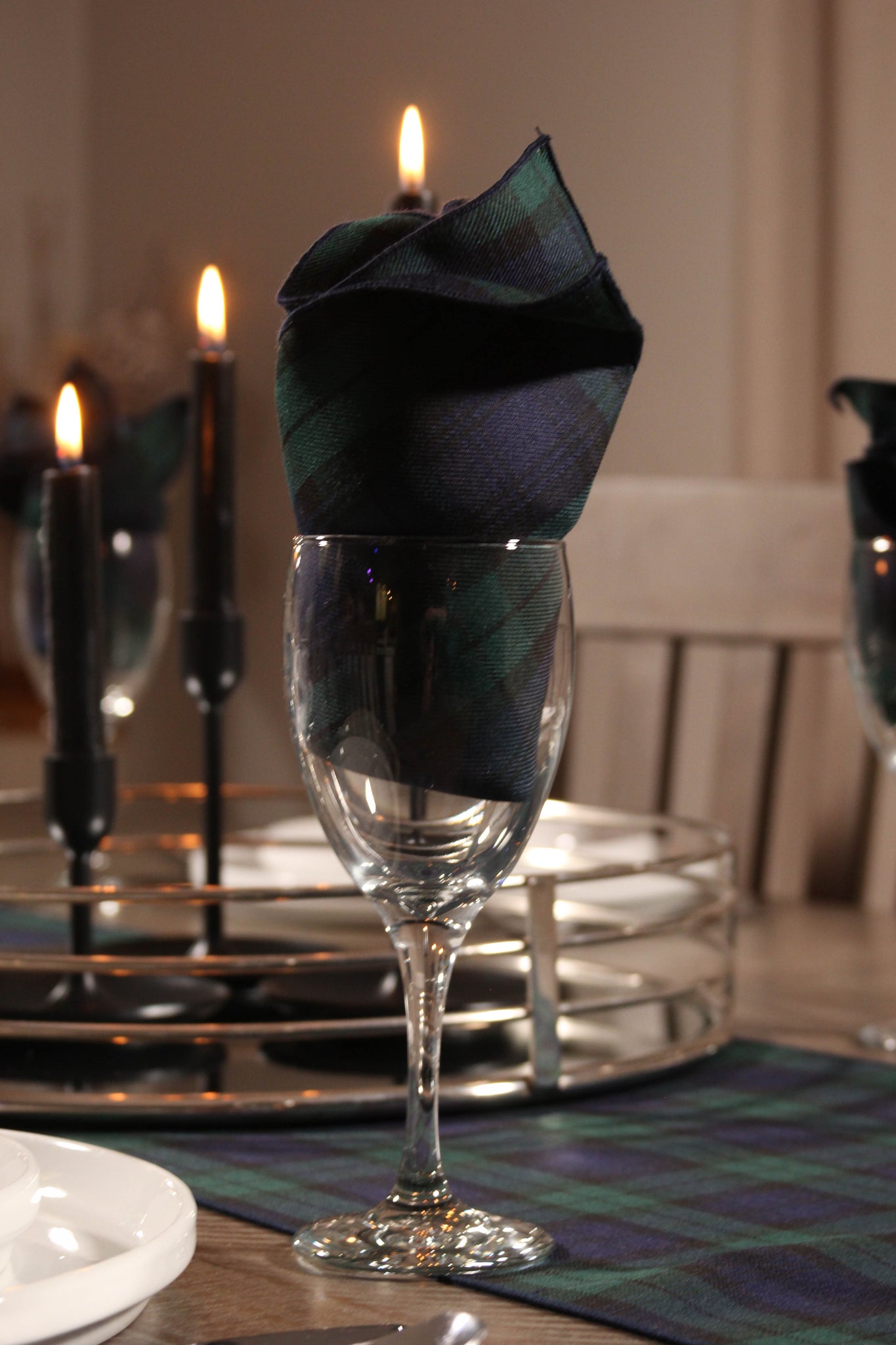 Black Watch plaid napkin displayed in a glass stemware, with a candle and matching table runner in the background.