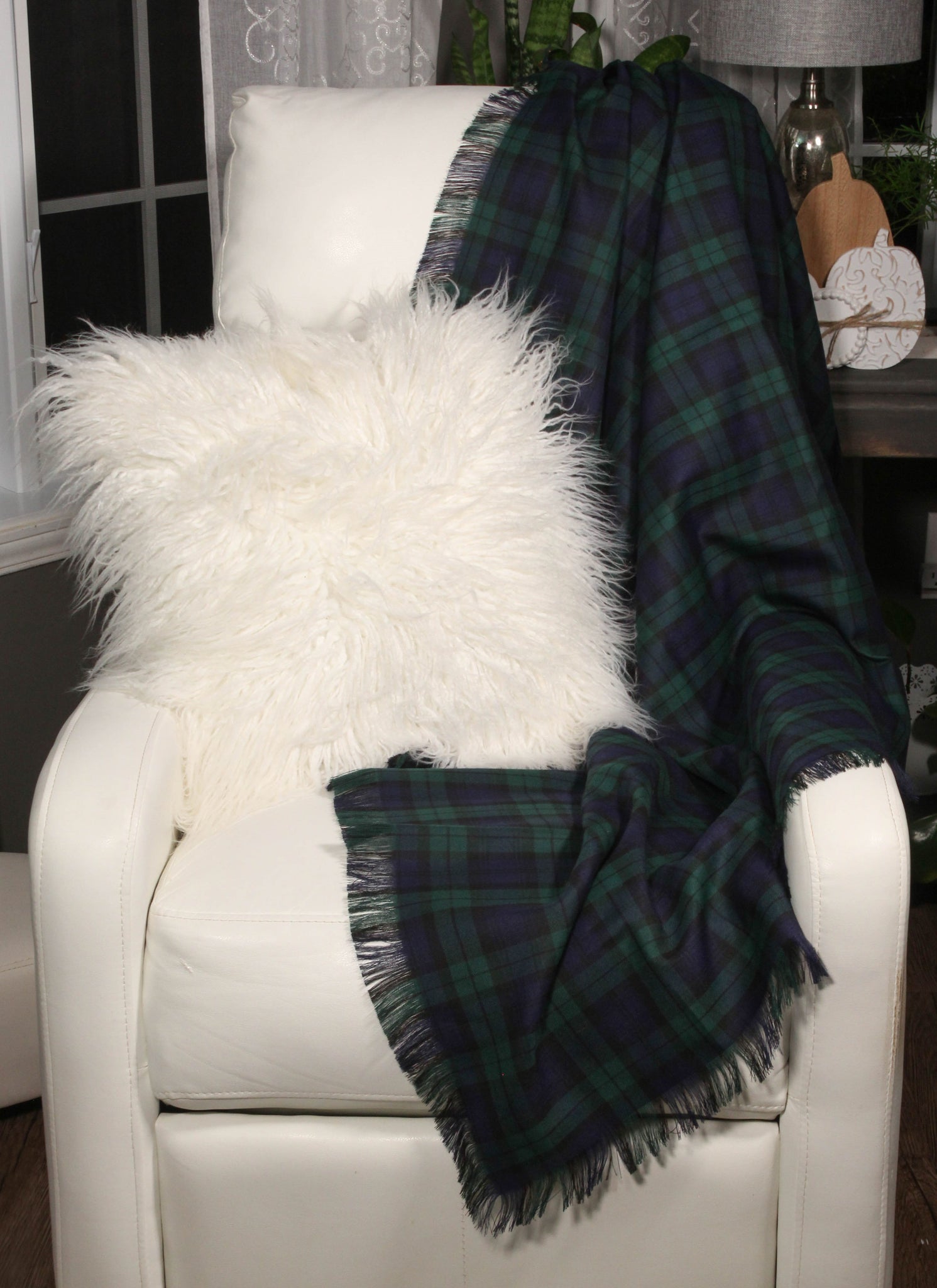 A Black Watch tartan throw with navy and green plaid pattern draped over a white sofa, complemented by a white furry pillow and a fringed throw pillow.