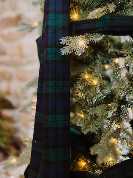 A close-up of a Black Watch tartan ribbon or fabric with a green and navy plaid pattern, draped over the branches of a flocked Christmas tree with warm glowing lights.