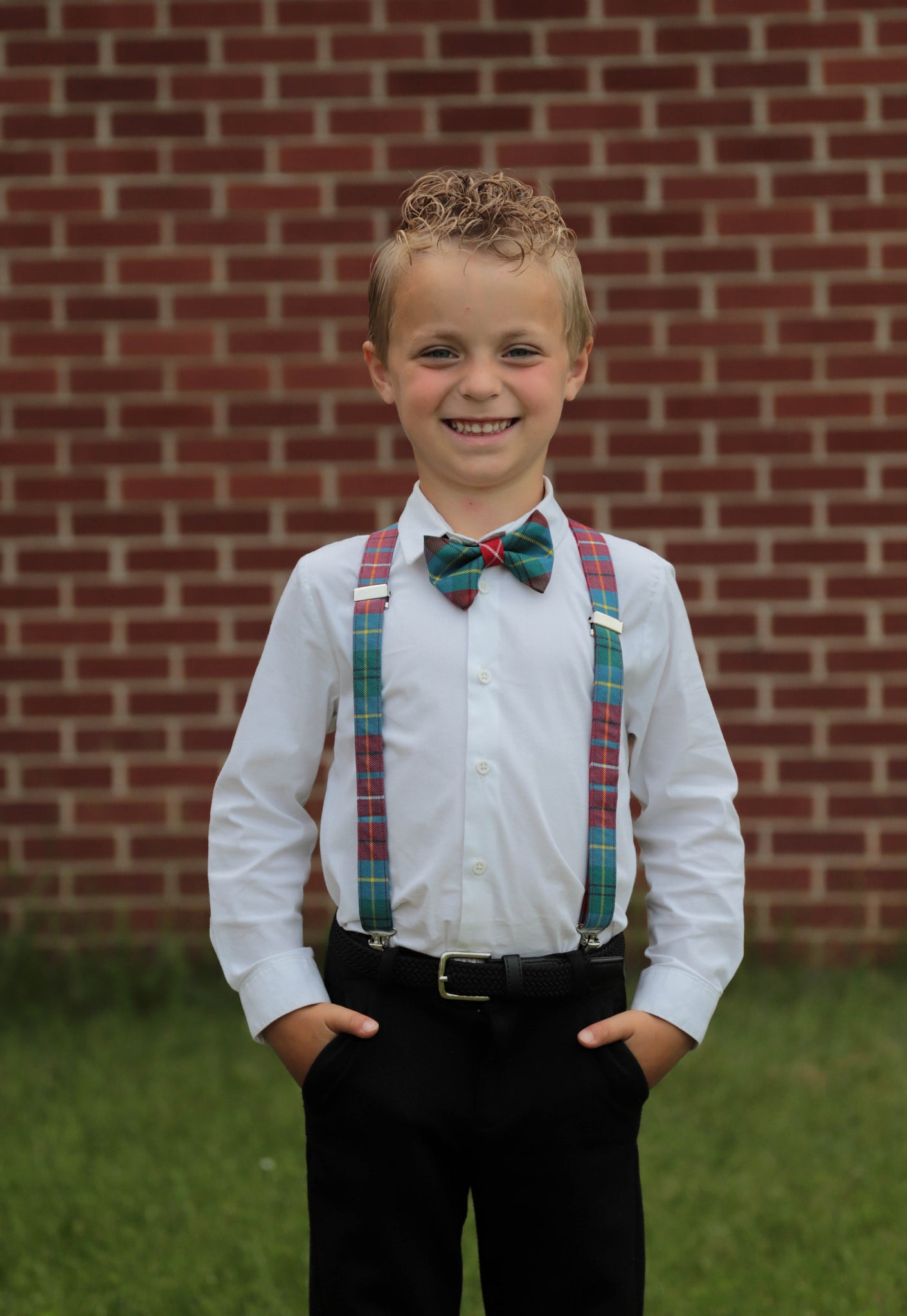 Young boy wearing a British Columbia tartan bow tie and matching suspenders in red, teal, white, and yellow plaid, paired with a white shirt and black dress pants.