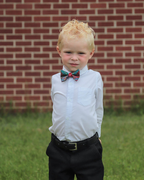 Young boy with blond curly hair in a white shirt and black pants with a bow tie standing in front of a brick wall.
