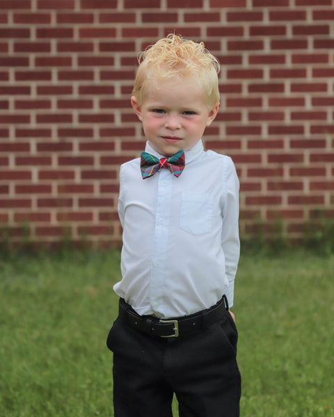 Young boy wearing a white shirt, black pants, and a red green white red and gold tartan bow tie standing in front of a brick wall.