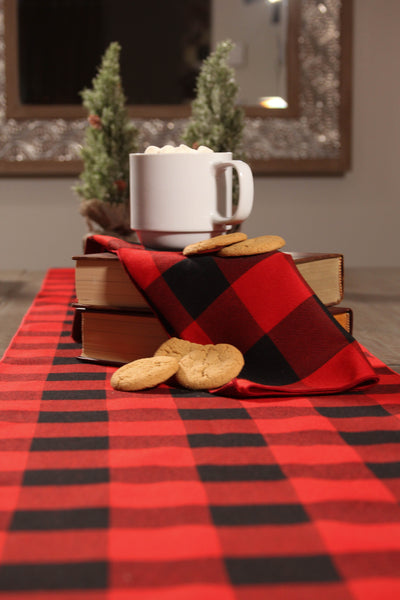 Close-up of a MacGregor red and black tartan napkin on a stack of books, with a cup of hot chocolate and cookies on top, and a matching table runner underneath.
