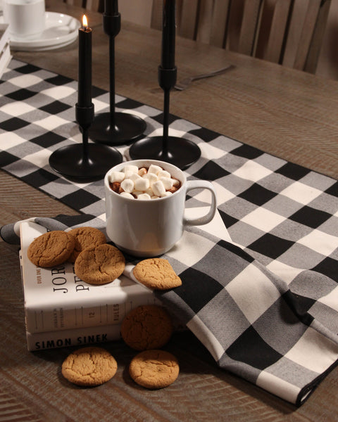 White mug with hot chocolate and marshmallows on a black and white checkered tablecloth with cookies and a book.