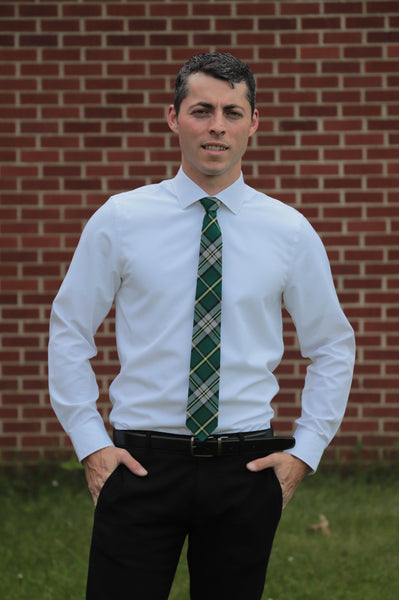 Man wearing a green, white, and yellow plaid tie with a white shirt and black pants standing in front of a brick wall.