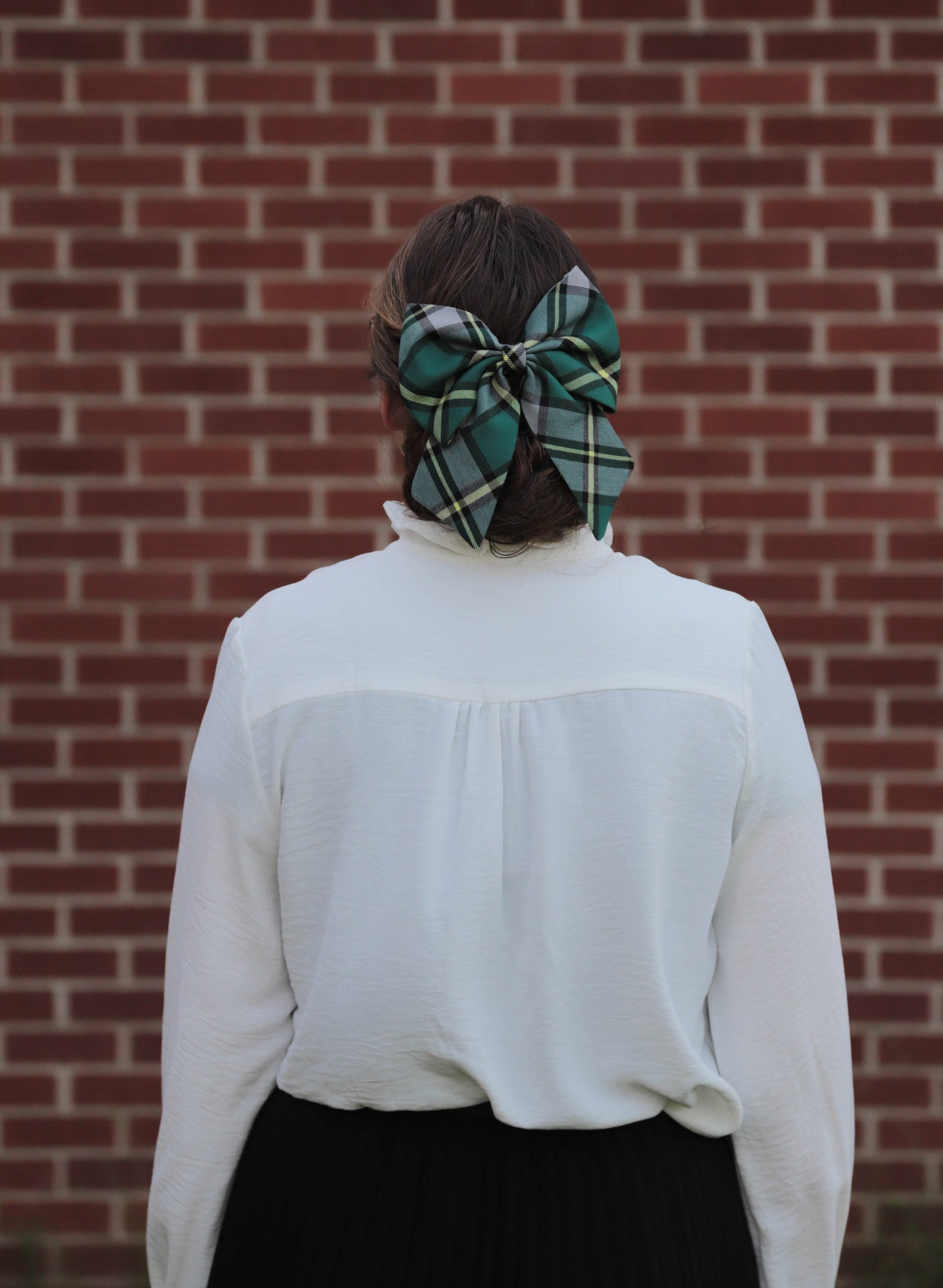 Woman wearing green plaid Cape Breton tartan hair bow, and white blouse in front of a brick wall