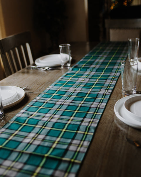 A green and white plaid Cape Breton tartan table runner laid down the center of a wooden dining table, surrounded by white plates and clear drinking glasses.