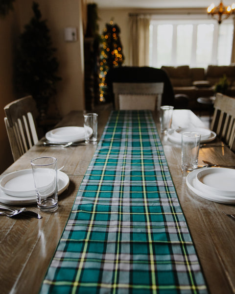 A green and teal plaid table runner laid down the center of a wooden dining table set with white plates and clear drinking glasses in a cozy, softly lit dining room.