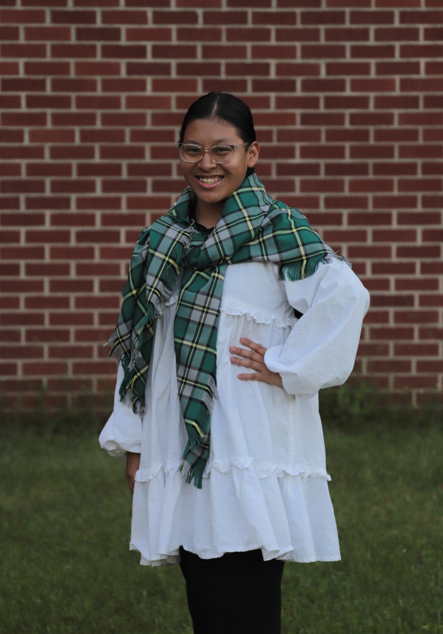 Person wearing a white blouse and Cape Breton tartan green, white, and yellow plaid blanket scarf in front of a brick wall.