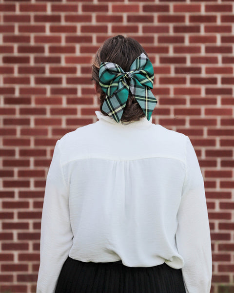 Woman wearing a green plaid Cape Breton tartan hair bow, white blouse, dark skirt against a brick wall