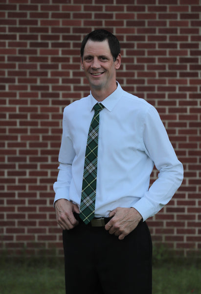 Man wearing a Cape Breton tartan plaid necktie with a white shirt and black dress pants standing in front of a red brick wall.