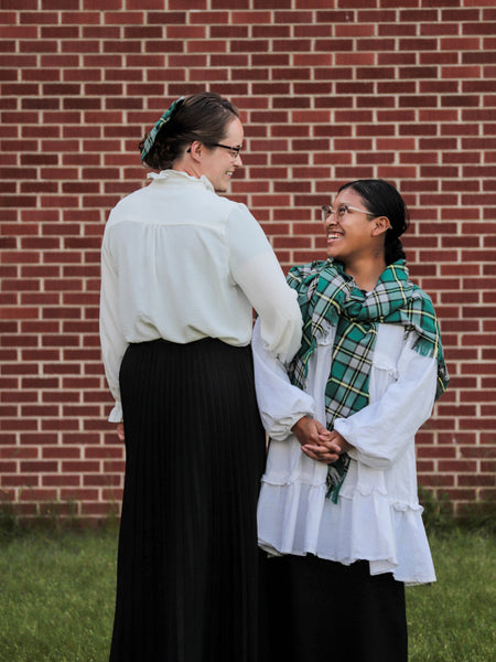 Two women wearing white shirts and black skirts with a cape breton tartan blanket scarf standing in front of a brick wall