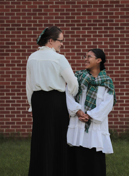 Two women with arms linked, wearing white blouses, green plaid oversized scarves, and matching hair bows in front of a brick wall.