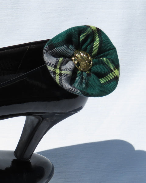 Green and gray plaid cape breton shoe clips with gold center on a black stand against a light blue background
