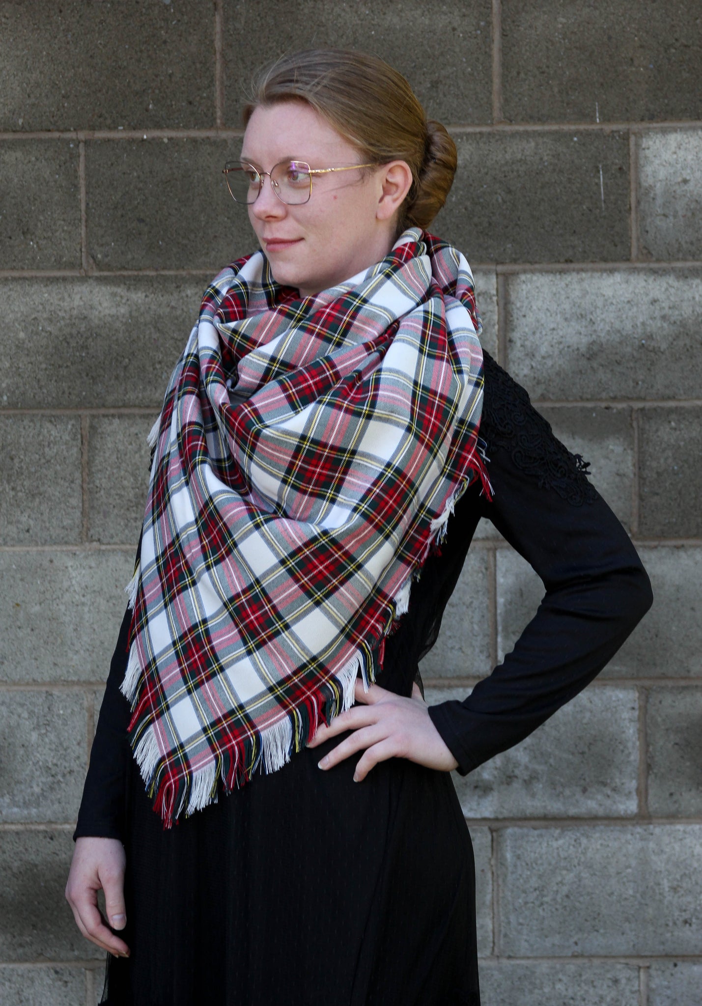 Woman wearing a Dress Stewart tartan white, red, and green plaid blanket scarf with fringed edges standing in front of a gray brick wall
