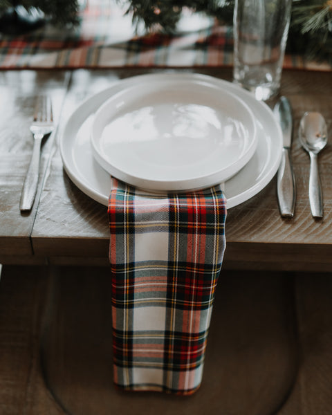 A close-up of a rustic wooden table set with white ceramic plates, silver cutlery, and a folded Dress Stewart tartan plaid napkin in red, green, black, and white tartan.