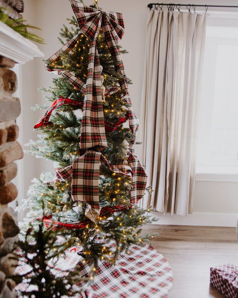 A decorated Christmas tree in a cozy living room, adorned with large red, green, and beige plaid bows, warm white string lights, and a matching Dress Stewart tartan plaid tree skirt. A stone fireplace, beige curtains, and a window are visible in the background.