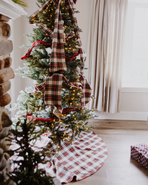 A Christmas tree decorated with red and beige plaid ribbons, warm white string lights, and a matching plaid tree skirt, set in a cozy room with a fireplace and window.