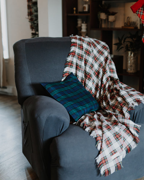 A cozy dark gray armchair in a living room setting, styled with a white plaid Dress Stewart tartan throw blanket and a blue-green plaid accent pillow.