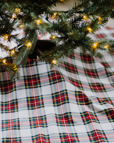 A close-up of a red, green, and white plaid Dress Stewart tartan Christmas tree skirt, draped under the branches of a decorated Christmas tree with warm string lights.